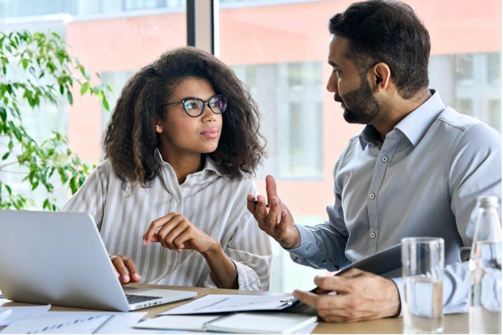 A woman with shoulder length brown hair speaking with a man with short black hair, in an office setting.